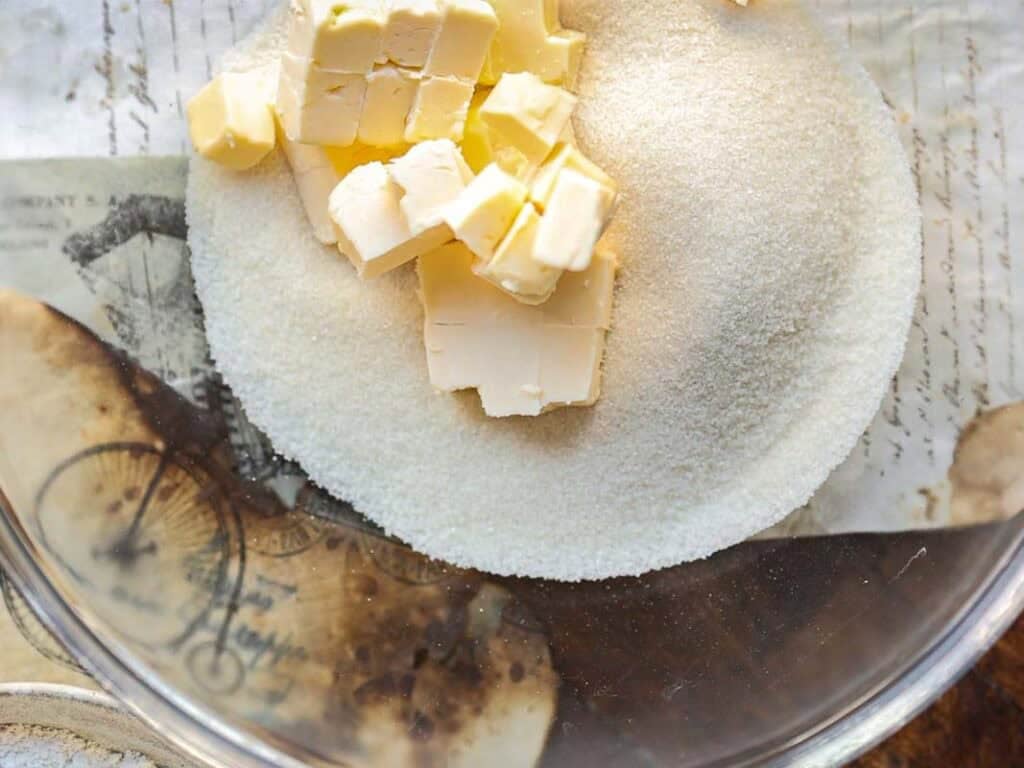 A glass bowl containing granulated sugar and cubed vegan butter sits on a vintage-style paper background.